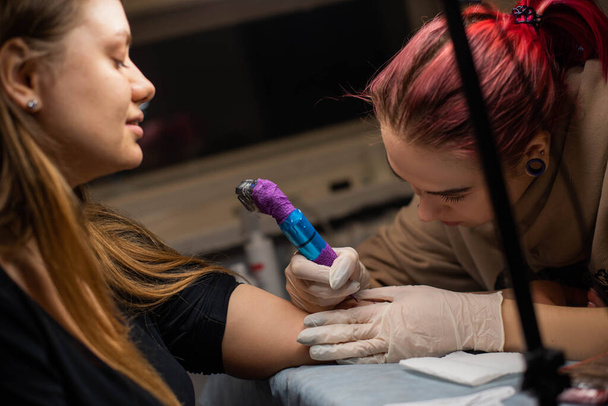 Encantadora chica con el pelo rosa y tatuajes llena un tatuaje en la mano de una mujer en un salón de tatuajes, pequeña empresa creativa y maestro del tatuaje. Hipster mujer en guantes dibuja un tatuaje - Foto, imagen