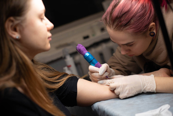 Bájos lány rózsaszín hajjal és tetoválásokkal egy tetoválást tesz egy nő kezére egy tetoválószalonban, kreatív kisvállalkozással és tetoválásmesterrel. Hipster nő kesztyűben rajzol egy tetoválást - Fotó, kép