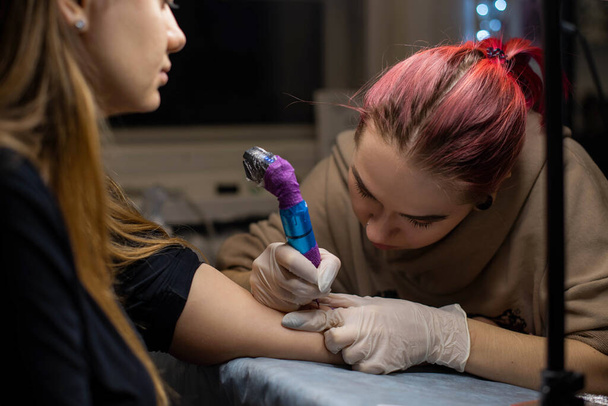 Okouzlující dívka s růžovými vlasy a tetování tetování na ženské ruce v tetovacím salónu, kreativní malé podnikání a tetování mistr. Hipster žena v rukavicích kreslí tetování - Fotografie, Obrázek