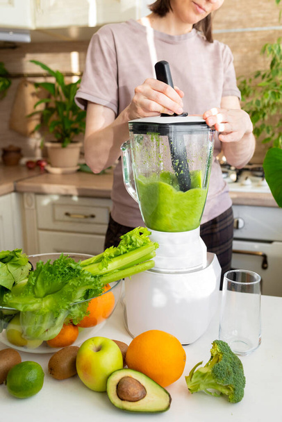 happy and cheerful caucasian woman in a t-shirt in her kitchen makes a green smoothie - Φωτογραφία, εικόνα