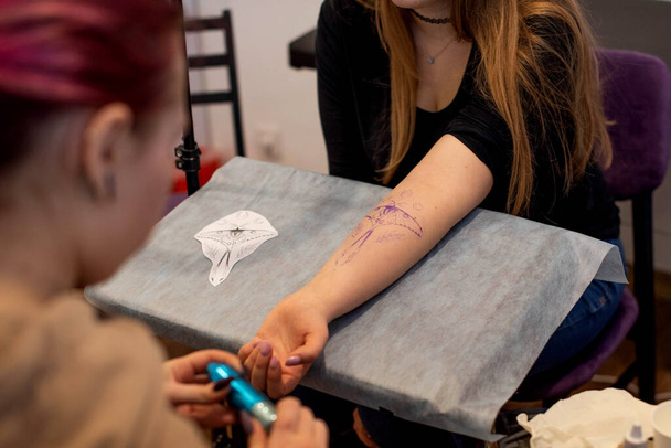 Encantadora chica con el pelo rosa y tatuajes llena un tatuaje en la mano de una mujer en un salón de tatuajes, pequeña empresa creativa y maestro del tatuaje. Hipster mujer en guantes dibuja un tatuaje - Foto, Imagen