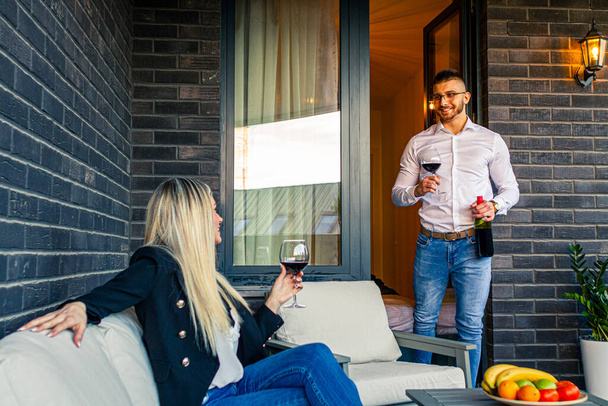 Genç bir çift gün batımında evlerinin terasında eğleniyor ve şarap içiyor.. - Fotoğraf, Görsel