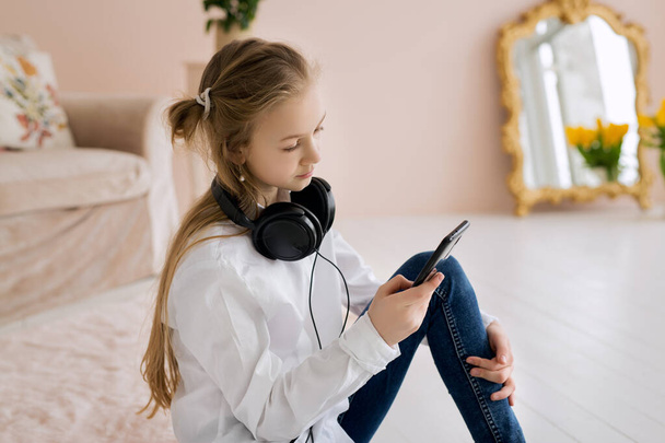 Дівчинка-підліток пише повідомлення на своєму смартфоні і слухає музику на навушниках. Великі навушники на шиї дівчини
. - Фото, зображення