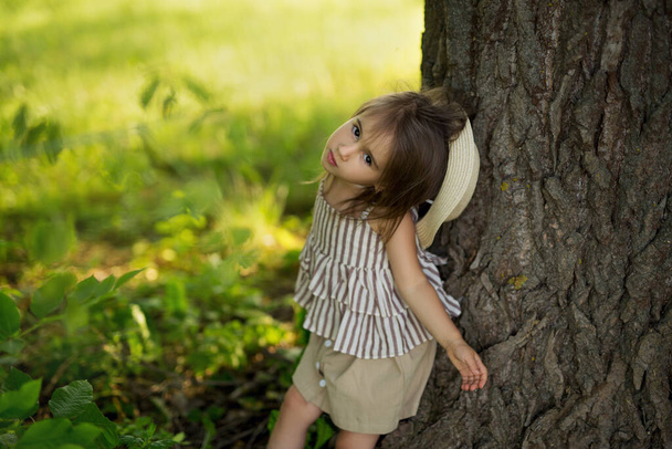 Pieni tyttö malli hattu poseeraa valokuvan valtava puunrunko puistossa. - Valokuva, kuva