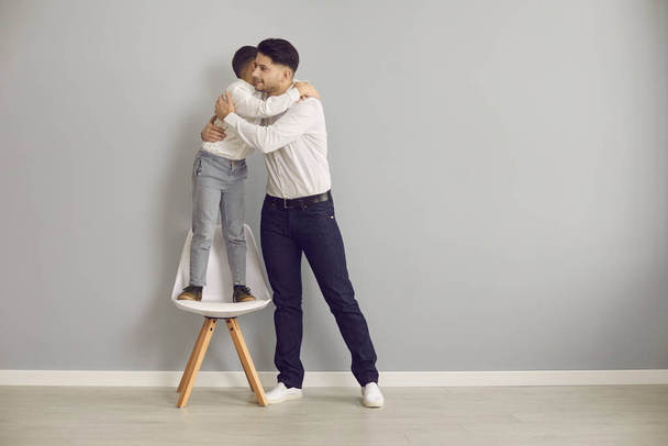 Loving father tightly hugging adorable son standing on chair studio portrait - Фото, зображення