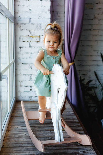 Een klein blond meisje heeft plezier en zwaait op een houten schommel paard in de buurt van het raam. - Foto, afbeelding