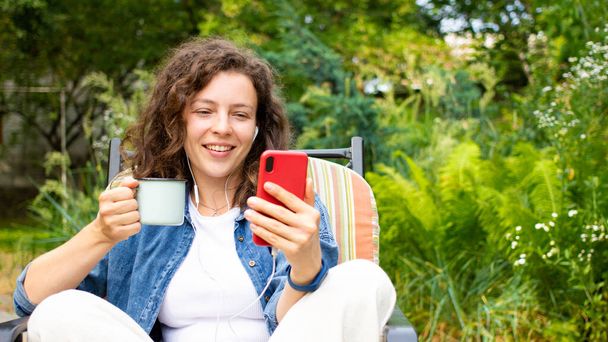 Krásná kudrnatá dívka pomocí smartphonu, držení šálek kávy, kontrola sociálních médií, chatování s přáteli, čtení zpráv, sledování online webinář, sedí na židli v zelené zahradě venku. - Fotografie, Obrázek