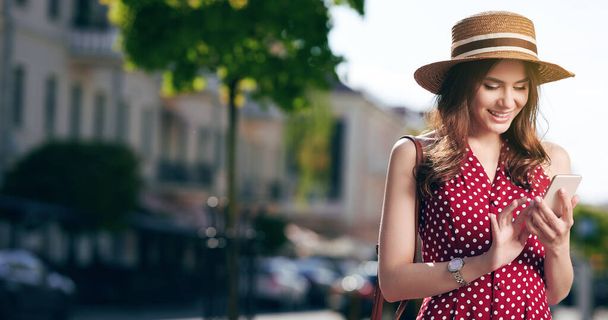 Caucasian female writes messages on her smartphone and walks in the summer city with a straw hat and a beautiful red dress with polka dots - Photo, Image