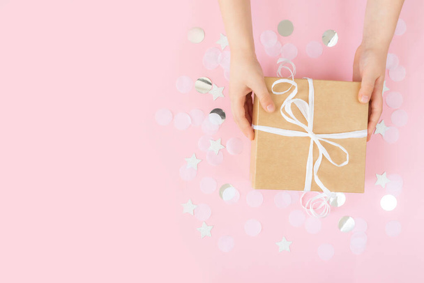 Flat lay with isolated adult hands holding craft paper gift box tied with ribbon, star and circle paper confetti or glitters on pink pastel background. High quality photo - Photo, Image