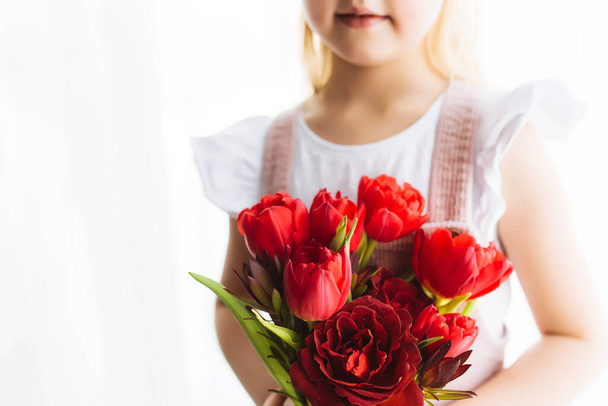 Small smiling girl holding bouquet of red tulip flowers. Concept for greeting card for Easter, Mother's day, International women's day, Saint Valentine's day. High quality photo - Foto, imagen