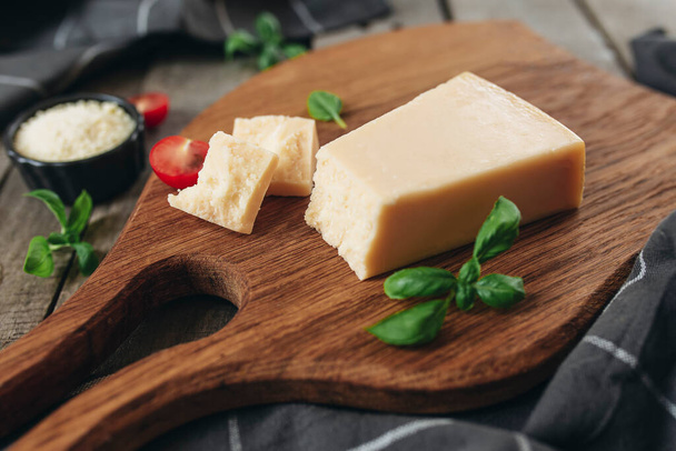 Italian cuisine concept. Cutting board, Parmesan Cheese chunks, grated cheese in small black bowl, cherry tomato cuts, basil leaves branches, kitchen towel on wooden background. Healthy eating concept - Valokuva, kuva