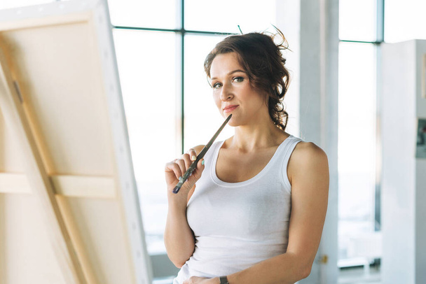 Pensando encantadora jovem artista em branco roupas casuais com pincéis de pintura na frente do cavalete na galeria de arte moderna - Foto, Imagem