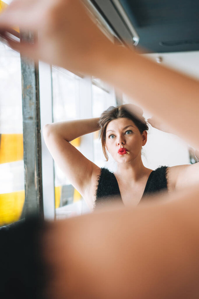 Belle jeune femme souriante plus corps de taille positive en robe de soirée noire faire coiffure près du miroir au studio de beauté - Photo, image