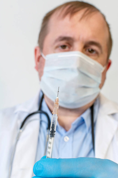 Mieslääkäri, jolla on stetoskoopilla varustettu suojaava naamio, pitää ruiskua, jossa on rokote. lähikuva. - Valokuva, kuva