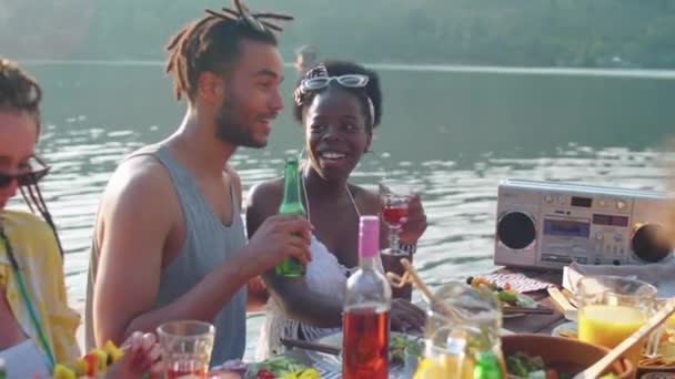 Schwenkbild der Gesellschaft junger multiethnischer Freunde, die am Esstisch auf der Seebrücke sitzen, lächeln und plaudern, während sie im Freien am malerischen See feiern - Filmmaterial, Video