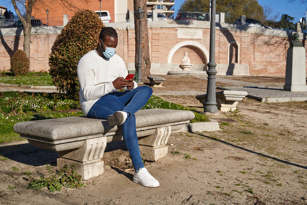 Молодий афроамериканець, який сидить на лавці в парку, посилає звістку. Латиноамериканець з протизаразною маскою. Концепція нової норми.  - Фото, зображення
