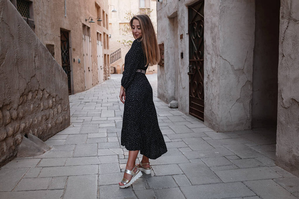 Ευτυχισμένη γυναίκα ταξιδιώτη φορώντας μαύρο φόρεμα περπατώντας στους δρόμους μιας παλιάς αραβικής πόλης ή χωριού στη μέση της ερήμου. Αντίληψη του τουρισμού και περιπέτειες στο Al Seef Dubai - Φωτογραφία, εικόνα
