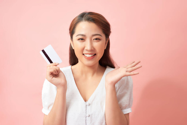 Bautiful happy young Asian woman showing credit card with open palm gesture isolated on light pink background - Fotoğraf, Görsel