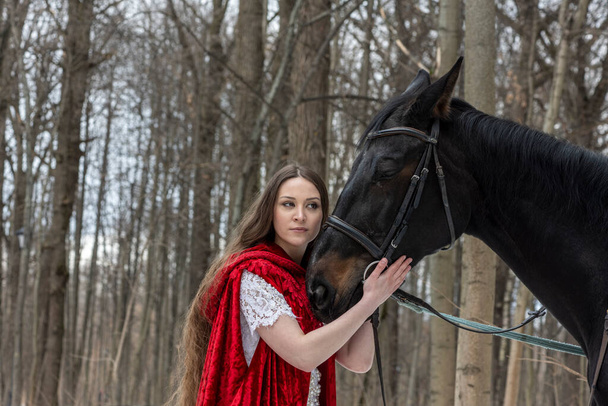 beautiful woman in white dress and red cape with black horse in spring forest  - Foto, immagini