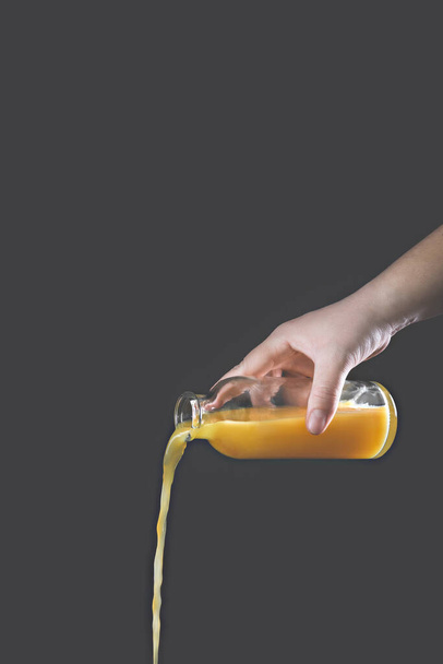 A woman's hand pours orange juice from a glass bottle on a gray background. Vertical orientation. Creative concept. - Foto, afbeelding