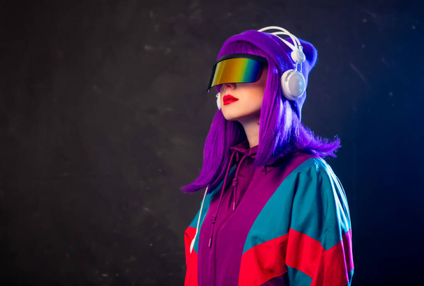 Stilvolles Mädchen mit Cyber-Punk-Brille und 80er-Jahre-Trainingsanzug mit Kopfhörern auf dunklem Hintergrund  - Foto, Bild