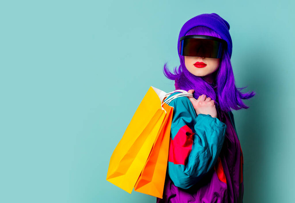 Stilvolles Mädchen in Cyber-Punk-Brille und 80er-Jahre-Trainingsanzug halten Einkaufstüten auf blauem Hintergrund - Foto, Bild