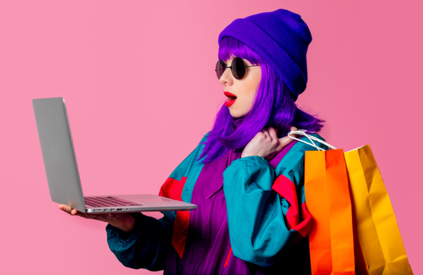 Stílusos lány 80-as évek melegítőben tartsa laptop és bevásárló táskák rózsaszín háttér - Fotó, kép