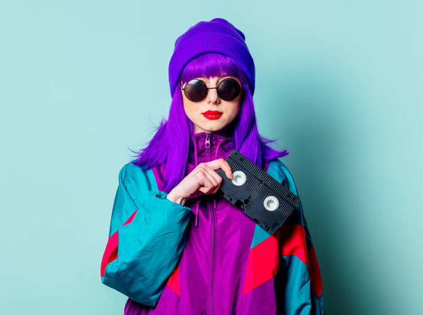 Стильна біла дівчина з фіолетовим волоссям, 80-х трек-костюм і сонцезахисні окуляри тримають відеокасету на синьому фоні
 - Фото, зображення