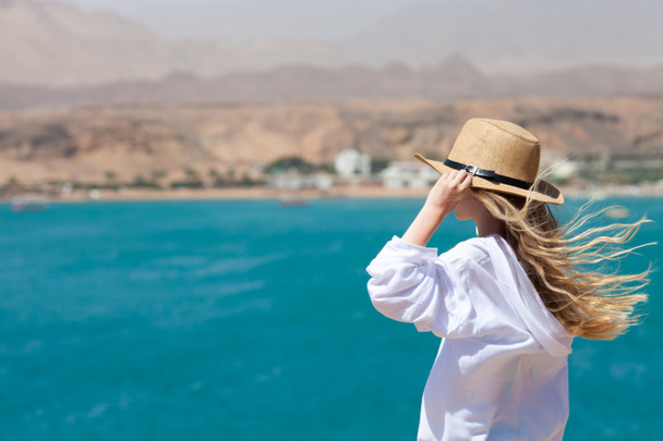 Blond girl with long hair blowing in the wind looking into the distance on the background of blue sea and mountains. Travel and tourism concept. Side view. - Photo, Image