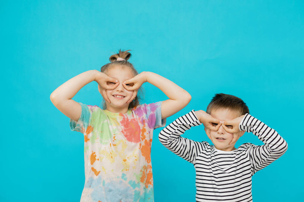 Aranyos kaukázusi gyerekek, egy lány és egy fiú fénymásoló szemüveget készítenek kék hátterű kezekből, fénymásolótérrel. Móka, mosoly, nevetés. Gyermek szórakozás és játékok. - Fotó, kép