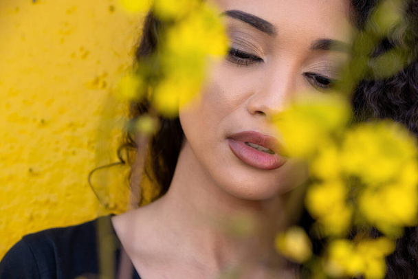 Beautiful woman with long curly hair standing behind delicate yellow blooming flowers - Foto, afbeelding