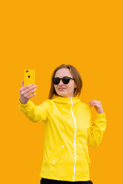 Een roodharig meisje in zonnebrillen en een regenjas staat met een smartphone in haar hand tegen een gele achtergrond en neemt selfie - Foto, afbeelding
