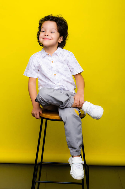 Photo of mixed race small kid, African American schoolboy, isolated over yellow background with copy space, a boy in street clothes sits, the curly-haired child looks away and smiles - Valokuva, kuva