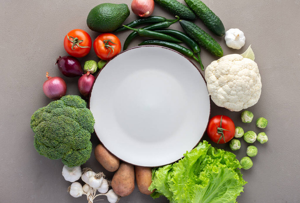 Vegetales orgánicos frescos de granja, concepto de alimentos saludables, verduras y champiñones alrededor de un plato redondo sobre fondo gris, vista superior, espacio de copia - Foto, Imagen