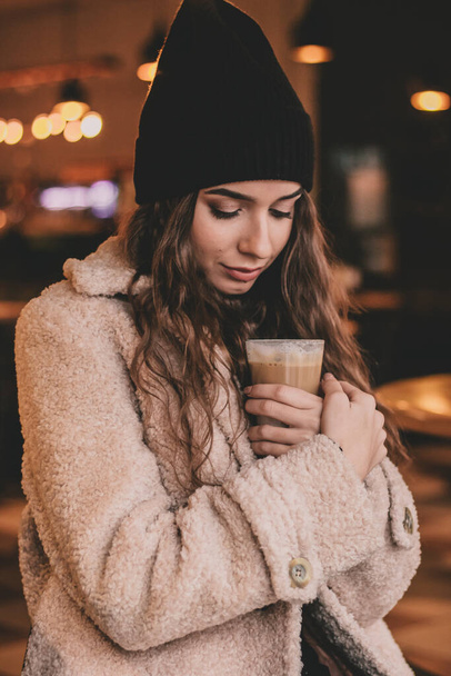 Una hermosa mujer de moda se sienta en una mesa en un restaurante sosteniendo un café con leche en sus manos y sonríe - Foto, imagen