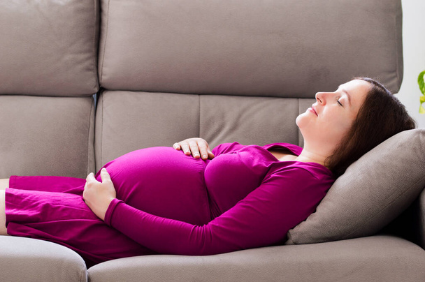 Portrait of a tired pregnant woman sleeping on a couch in the living room at home - 写真・画像