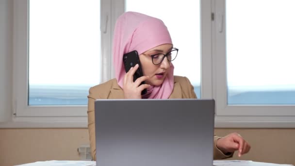 Woman in hijab examines documents and works on laptop while talking on the phone - Footage, Video