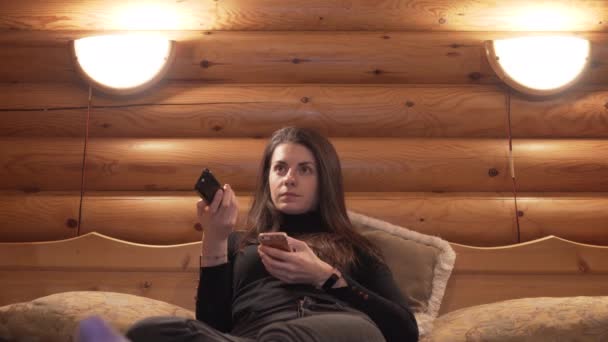 Mujer joven cambiando los canales de televisión de la cama grande en cabaña de madera de invierno - Imágenes, Vídeo