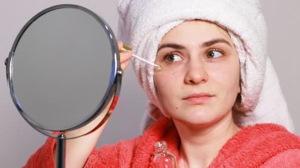 Kaunis nuori nainen vaaleanpunaisessa kylpytakissa, jolla on valkoinen pyyhe päässään, levittää öljyä vitamiineilla kasvoilleen ravitsemaan ja kosteuttamaan ihoa. Ihonhoito kosmetiikassa - Materiaali, video