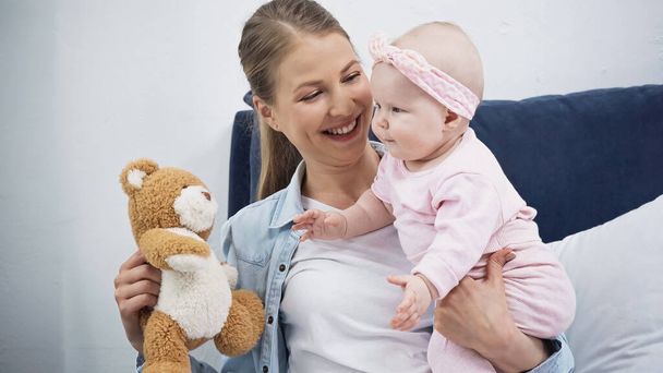 cheerful woman holding soft toy near infant daughter in headband with bow - Foto, afbeelding