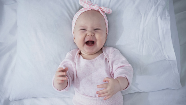 top view of upset baby girl in headband with bow crying while lying on bed - Foto, imagen