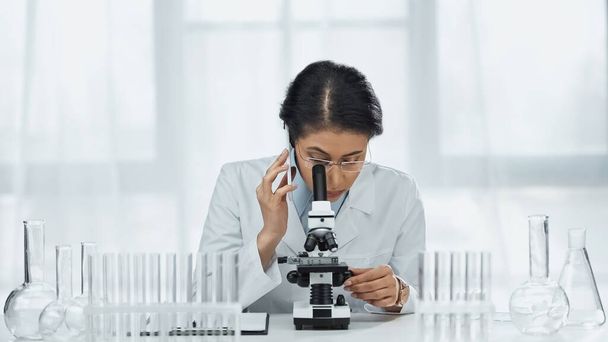 científico afroamericano en gafas hablando en smartphone mientras mira a través del microscopio en el laboratorio  - Foto, Imagen