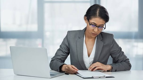 african american businesswoman in glasses looking at papers on clipboard near laptop - Fotografie, Obrázek