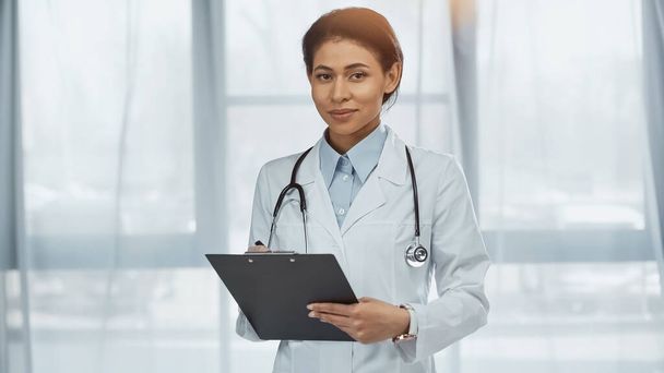 американский врач в белом халате со стетоскопом, держащим планшет в клинике  - Фото, изображение