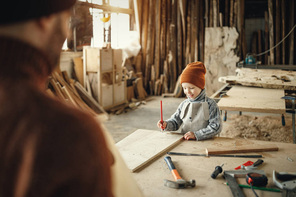 Happy little kid with pencil in hand making marks on wooden plank while assisting father during work in carpentry workshop - Photo, Image