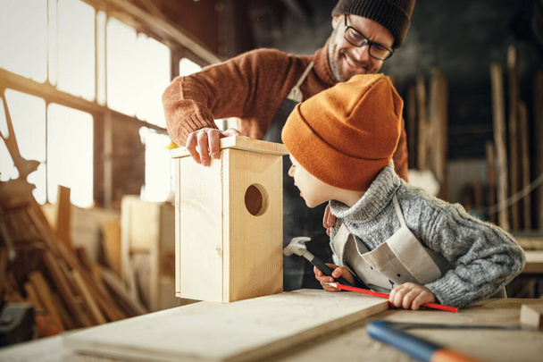 Cheerful adult bearded man and little son in aprons assembling wooden bird house together while working in carpentry workshop - Fotó, kép