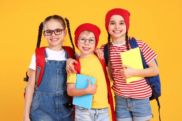 Compañeros de clase primaria optimistas en ropa casual con mochilas mirando a la cámara mientras están de pie juntos contra el fondo amarillo - Foto, Imagen