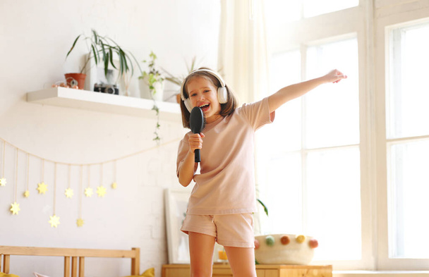 I am super star. Happy preschool child wearing pajama holding hair brush as microphone and singing her favourite song while standing on the bed at home, cute little girl pretending to be famous singer - Photo, Image