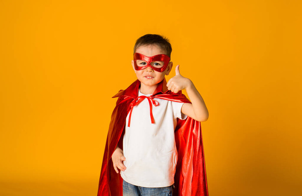retrato de un niño pequeño con un disfraz de héroe con una máscara roja y una capa muestra la clase con su mano sobre un fondo amarillo con espacio para el texto - Foto, imagen