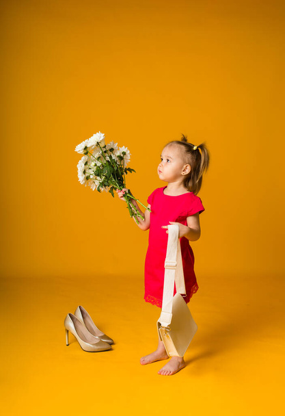 little girl with ponytails in a red dress stands sideways with a bouquet of white flowers on a yellow background with space for text - Zdjęcie, obraz